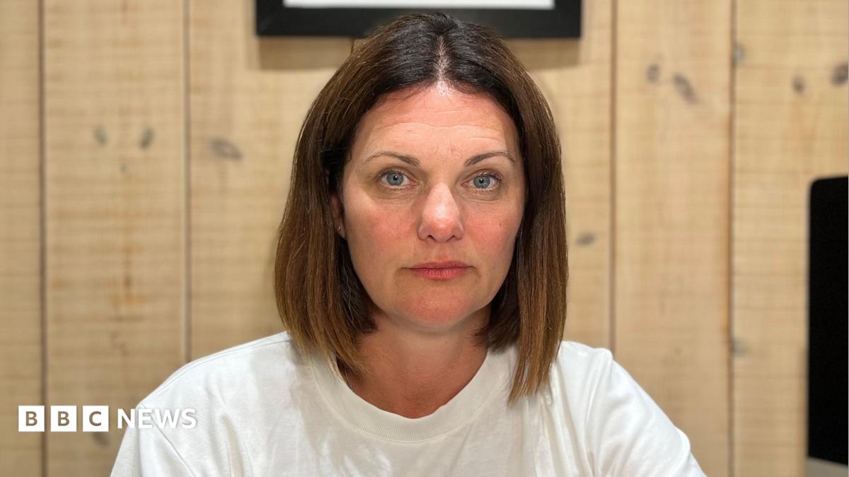 A woman with dark hair and blue eyes in a plain white T-shirt sits at a desk in a wood-panelled home office, facing the camera. A computer monitor, notebook, water bottle, phone and glasses are visible on the desk, with framed artwork hanging on the wall behind.