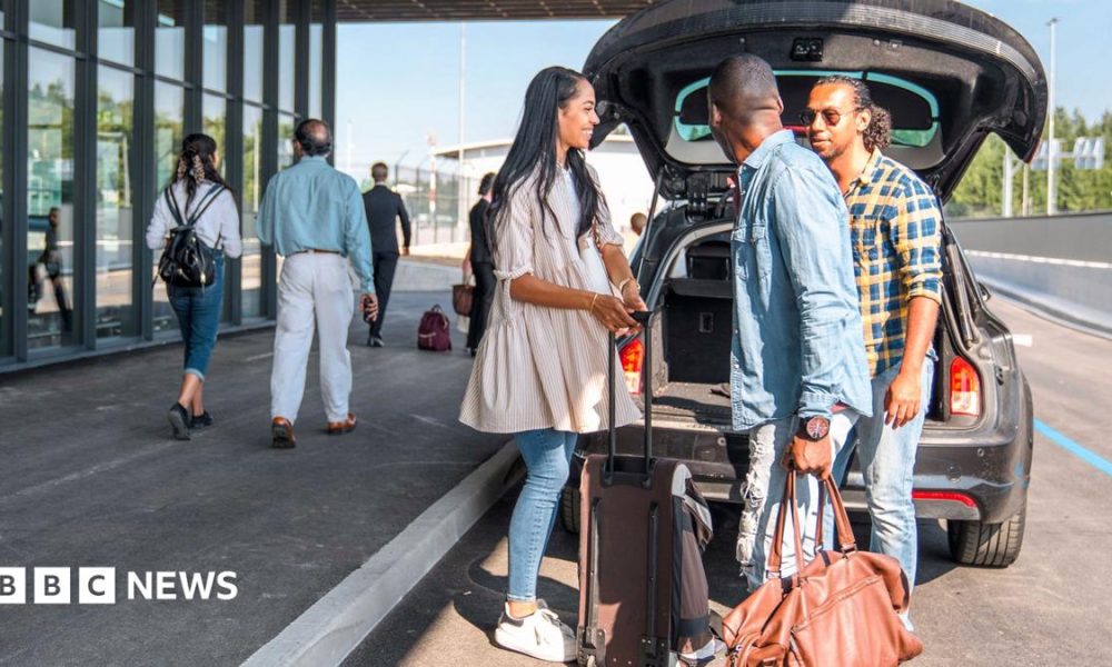 A sign outside Gatwick airport reads 'check-in & bag drop; north terminal Gatwick', while people walk past wheeling baggage along the concrete floor, image taken in 2024.