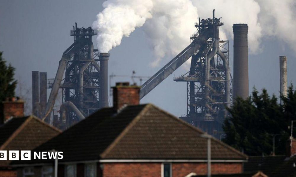 A British Steel plant, with steam billowing out of it, behind a row of terrace houses