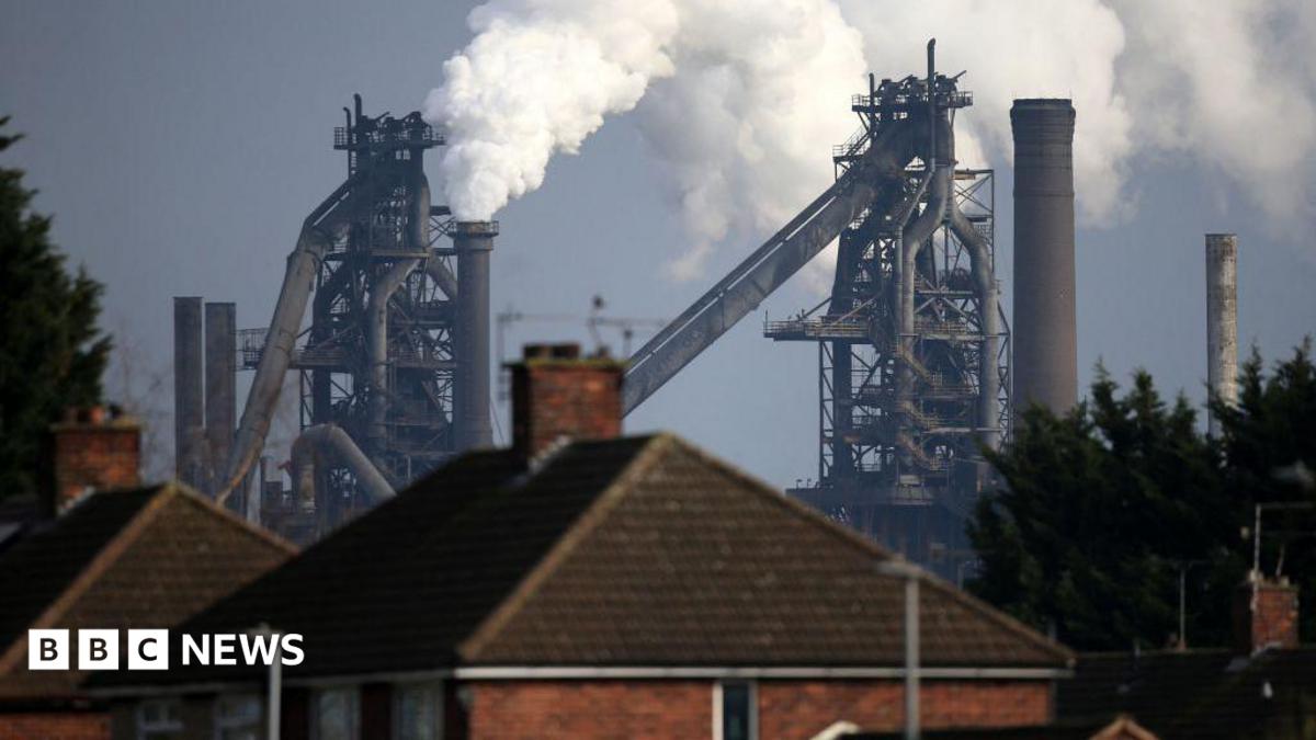 A British Steel plant, with steam billowing out of it, behind a row of terrace houses