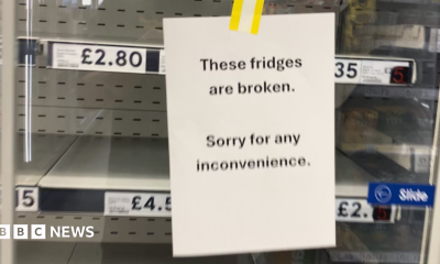 A sign saying 'these fridges are broken, sorry for any inconvenience'. There are empty fridge shelves behind it.