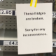 A sign saying 'these fridges are broken, sorry for any inconvenience'. There are empty fridge shelves behind it.