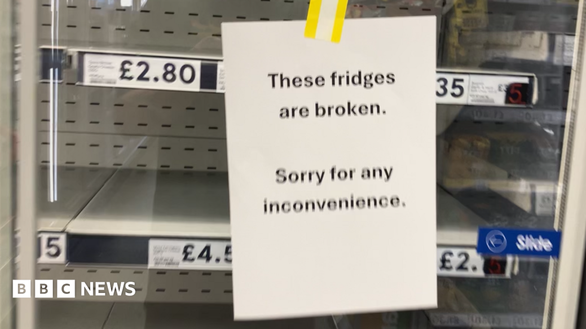A sign saying 'these fridges are broken, sorry for any inconvenience'. There are empty fridge shelves behind it.