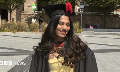 Person standing in a paved public square wearing a graduation gown and mortarboard with a tassel. A yellow dress is visible beneath the gown. Trees, ornamental grasses and stone buildings surround the square, with several people seated or walking in the background.