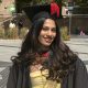 Person standing in a paved public square wearing a graduation gown and mortarboard with a tassel. A yellow dress is visible beneath the gown. Trees, ornamental grasses and stone buildings surround the square, with several people seated or walking in the background.