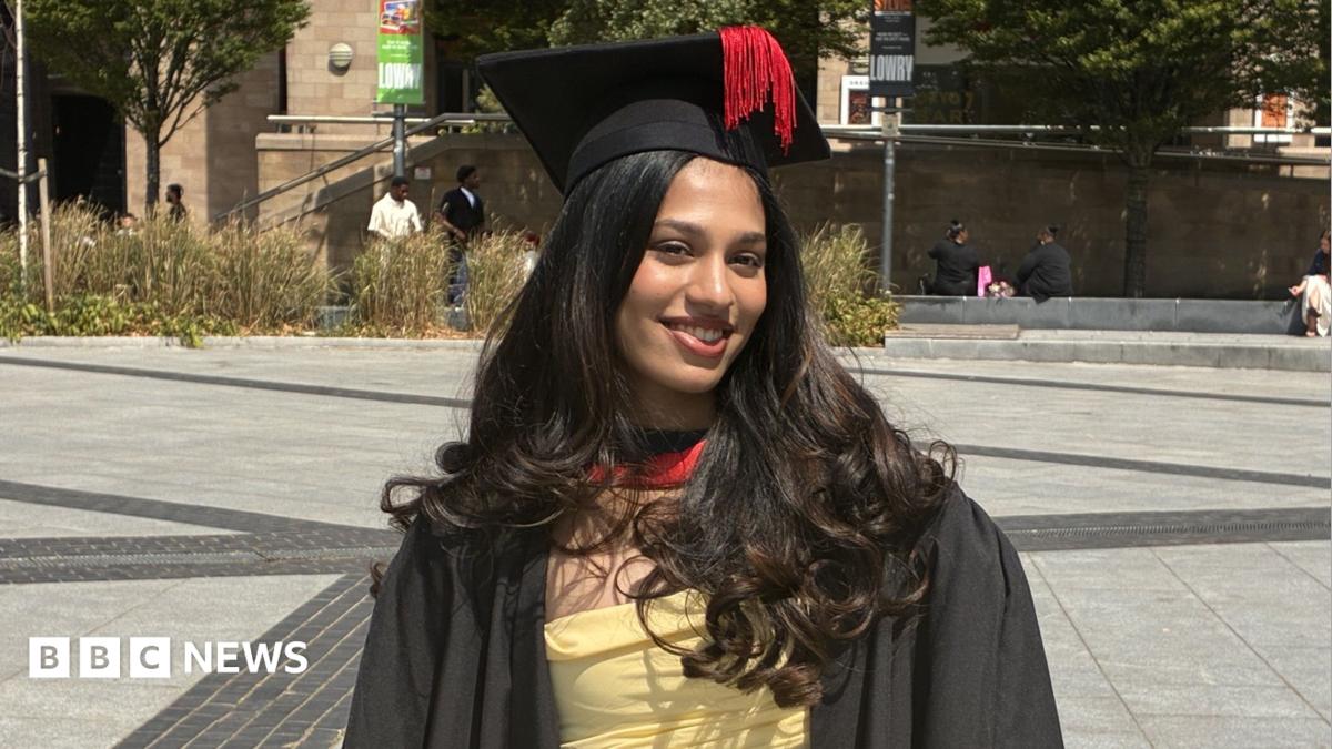Person standing in a paved public square wearing a graduation gown and mortarboard with a tassel. A yellow dress is visible beneath the gown. Trees, ornamental grasses and stone buildings surround the square, with several people seated or walking in the background.