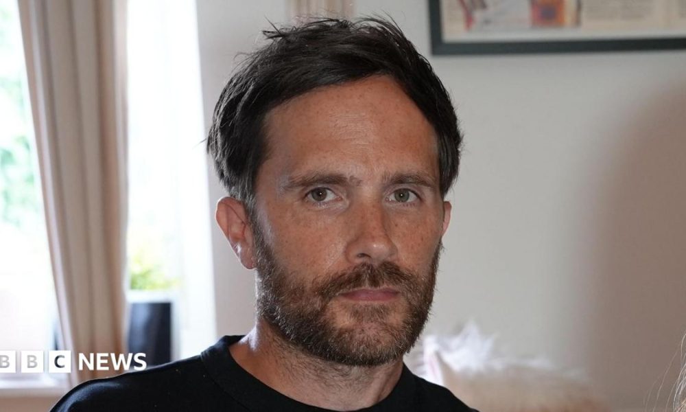 Craig Bowe sitting in a living room. He has dark hair and a brown beard. He looks serious in his expression while wearing a black T-shirt.
