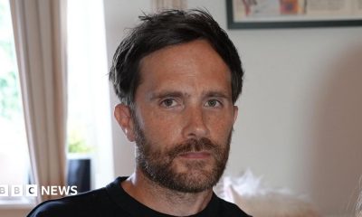 Craig Bowe sitting in a living room. He has dark hair and a brown beard. He looks serious in his expression while wearing a black T-shirt.