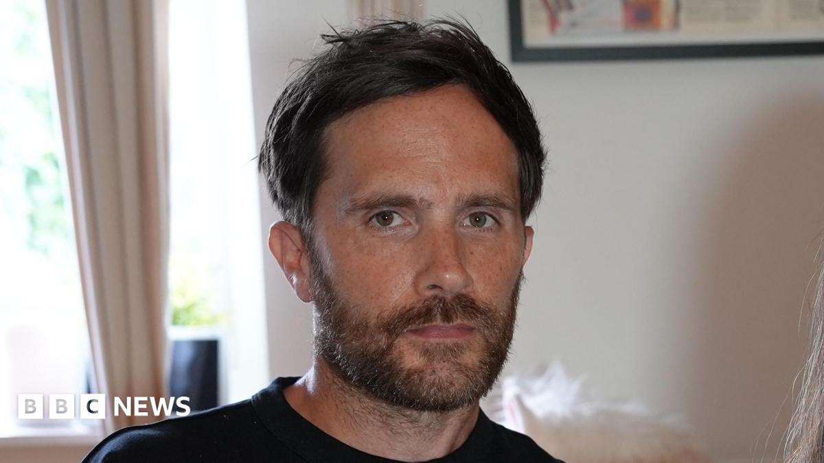 Craig Bowe sitting in a living room. He has dark hair and a brown beard. He looks serious in his expression while wearing a black T-shirt.