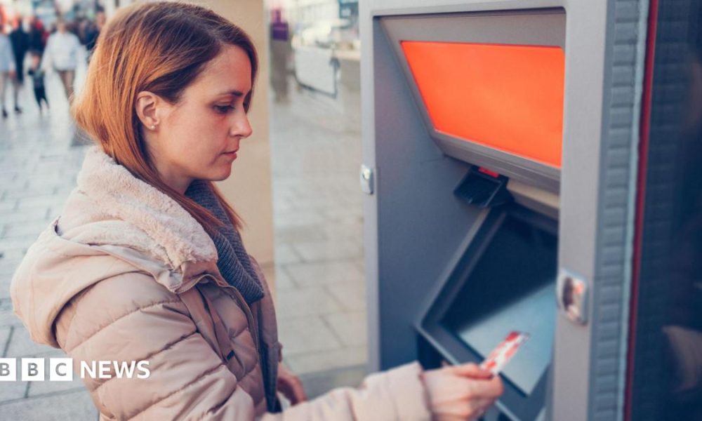 Woman wearing a beige puffer coat holds a debit card while standing in front of an ATM.