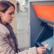 Woman wearing a beige puffer coat holds a debit card while standing in front of an ATM.