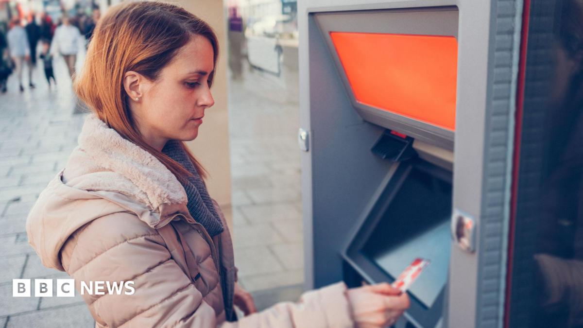 Woman wearing a beige puffer coat holds a debit card while standing in front of an ATM.