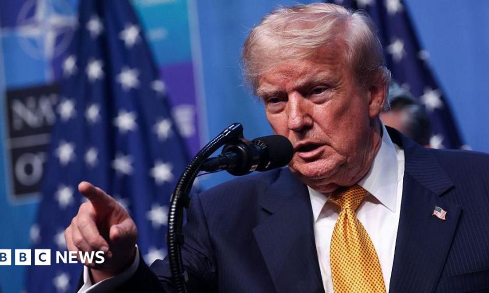 US President Donald Trump wearing a yellow tie, white shirt, blue suit and American flag pin. He stands at a microphone and is pointing, his mouth open.
