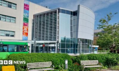 A large glass-fronted building with two park benches at its front. There is a green bus driving pass the entrance of the hospital.