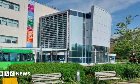 A large glass-fronted building with two park benches at its front. There is a green bus driving pass the entrance of the hospital.