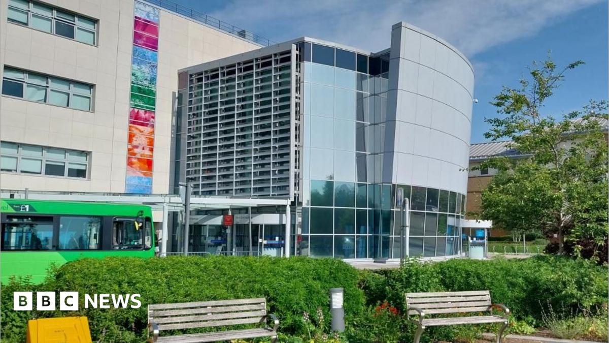 A large glass-fronted building with two park benches at its front. There is a green bus driving pass the entrance of the hospital.
