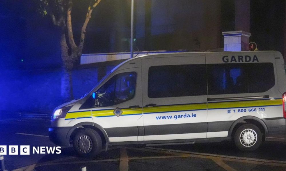 A white Garda van with a yellow stripe along its side pulls out onto a main road on a dark night.