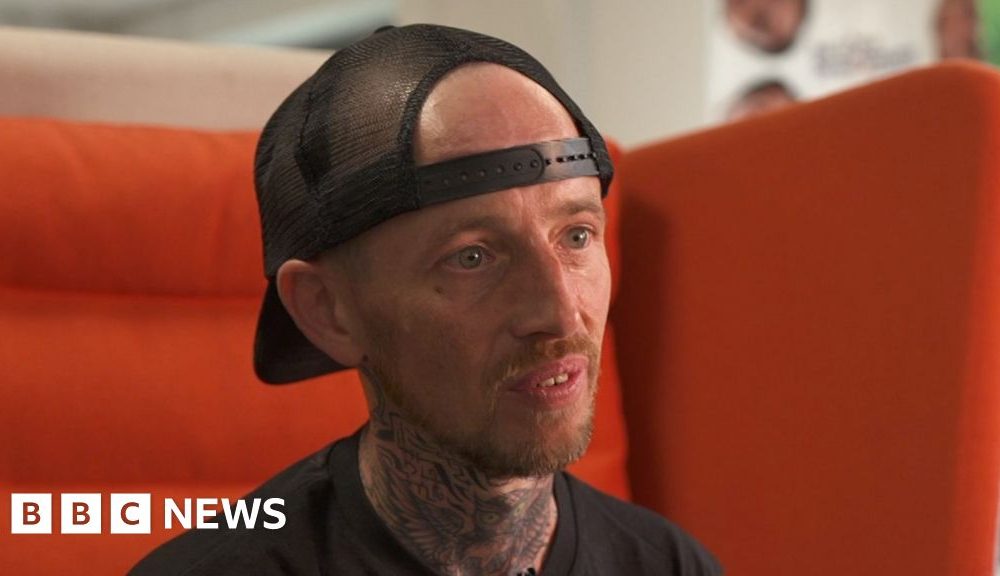 Steven, a man in a black t shirt and black snapback cap sat on a large orange chair.  He has a short red haired goatee.