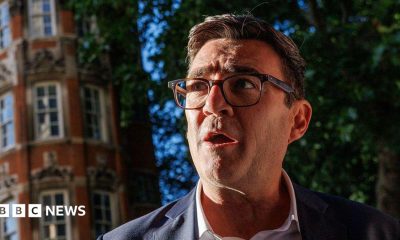 Andy Burnham, wearing a suit, white collared shirt and glasses. A red-brick building and a tree are in the background.