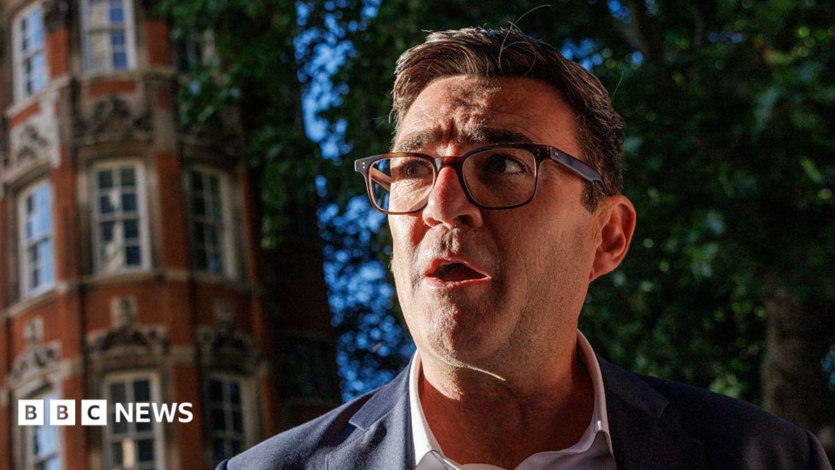 Andy Burnham, wearing a suit, white collared shirt and glasses. A red-brick building and a tree are in the background.