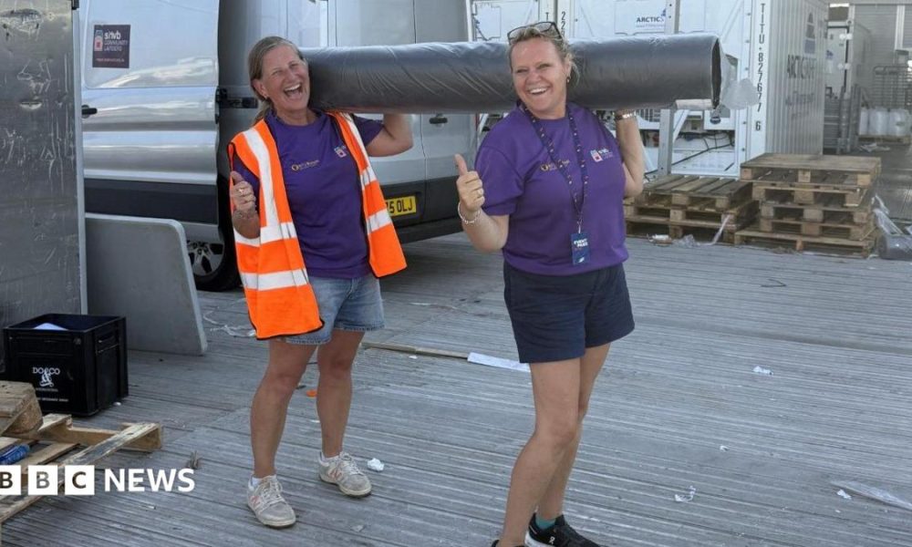 Two women, both holding their thumbs up, holding a carpet roll, both wearing shorts, and a purple T-shirt. The one on the left has on a high-vis jacket. A silver van is behind them and some wooden pallets.