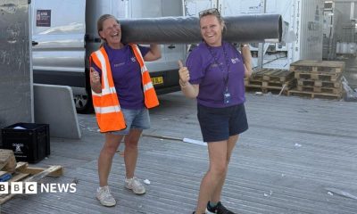Two women, both holding their thumbs up, holding a carpet roll, both wearing shorts, and a purple T-shirt. The one on the left has on a high-vis jacket. A silver van is behind them and some wooden pallets.