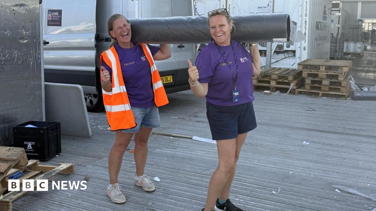 Two women, both holding their thumbs up, holding a carpet roll, both wearing shorts, and a purple T-shirt. The one on the left has on a high-vis jacket. A silver van is behind them and some wooden pallets.