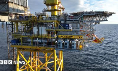 Offshore oil and gas platform marked “JACKDAW 30/2a” standing on yellow support legs above the sea, with industrial equipment, pipework and a crane visible under a partly cloudy sky.