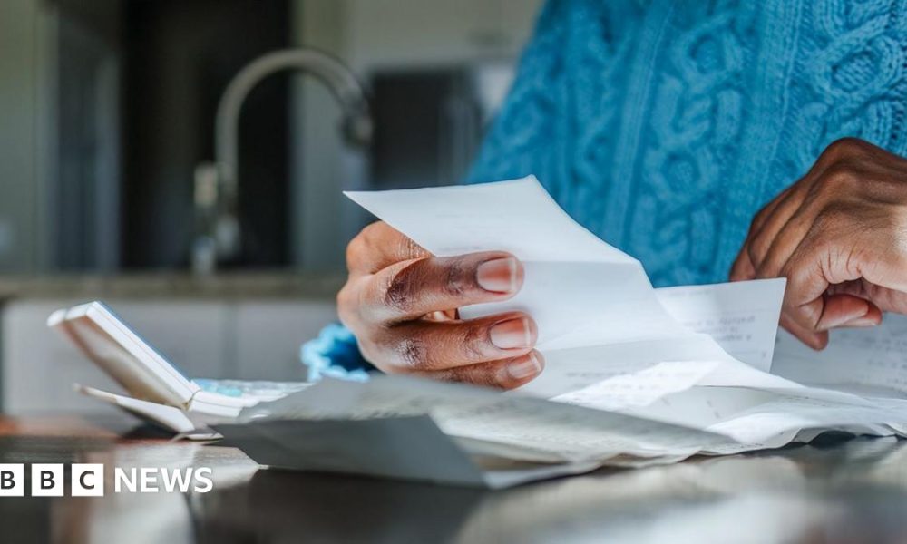 Close up shot of a person looking at bills and receipts on a table next to a calculator in their kitchen. They are wearing a blue jumper and the kitchen sink is in the background.