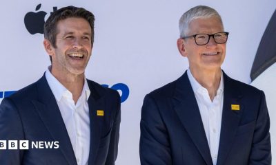 Apple's incoming chief executive John Ternus standing next to his predecessor Tim Cook. Both are smiling for cameras at an Apple event and wearing an almost identical blue blazer and white collared shirt.