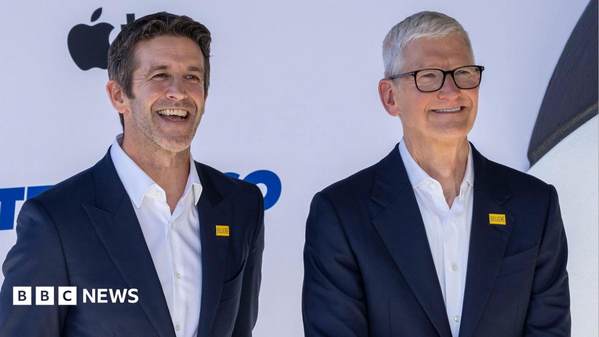Apple's incoming chief executive John Ternus standing next to his predecessor Tim Cook. Both are smiling for cameras at an Apple event and wearing an almost identical blue blazer and white collared shirt.