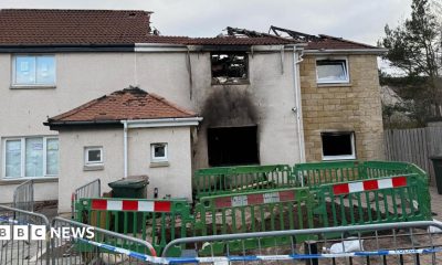 A house damaged by fire. The house is made of white and yellow stone. The roof has caved in and is black with fire damage. In front of it is a green barrier and in front of that is a silver barrier with white and blue police tape attached.