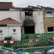 A house damaged by fire. The house is made of white and yellow stone. The roof has caved in and is black with fire damage. In front of it is a green barrier and in front of that is a silver barrier with white and blue police tape attached.