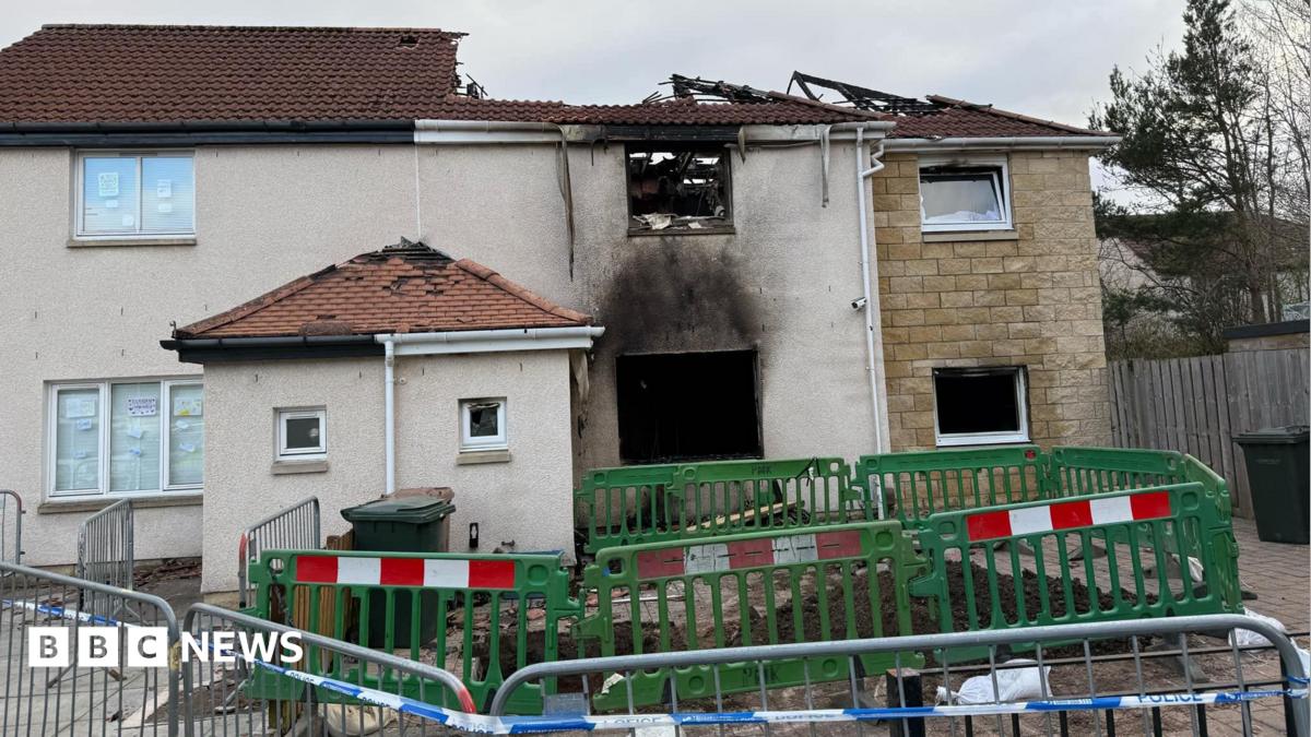 A house damaged by fire. The house is made of white and yellow stone. The roof has caved in and is black with fire damage. In front of it is a green barrier and in front of that is a silver barrier with white and blue police tape attached.