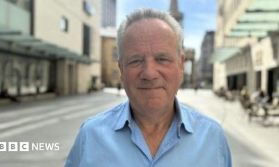Chris wears a light blue collared shirt and has a mild smile on his face, while standing outside on a sunny day surounded by buildings.