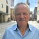 Chris wears a light blue collared shirt and has a mild smile on his face, while standing outside on a sunny day surounded by buildings.