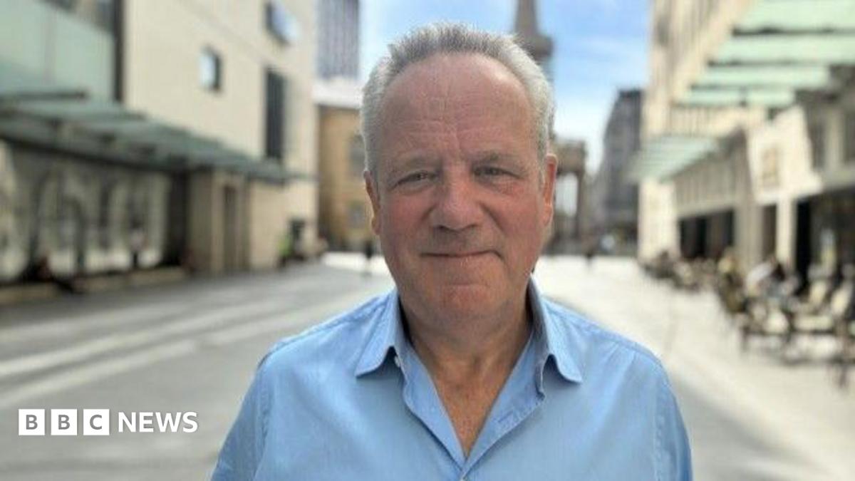 Chris wears a light blue collared shirt and has a mild smile on his face, while standing outside on a sunny day surounded by buildings.