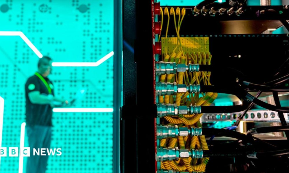 A man standing against a bright green background in a data centre - in the foreground is the back of a large computer with lots of yellow and black wires.