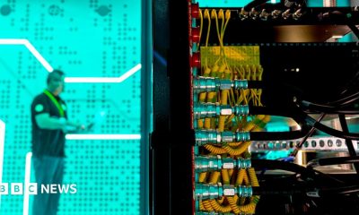 A man standing against a bright green background in a data centre - in the foreground is the back of a large computer with lots of yellow and black wires.