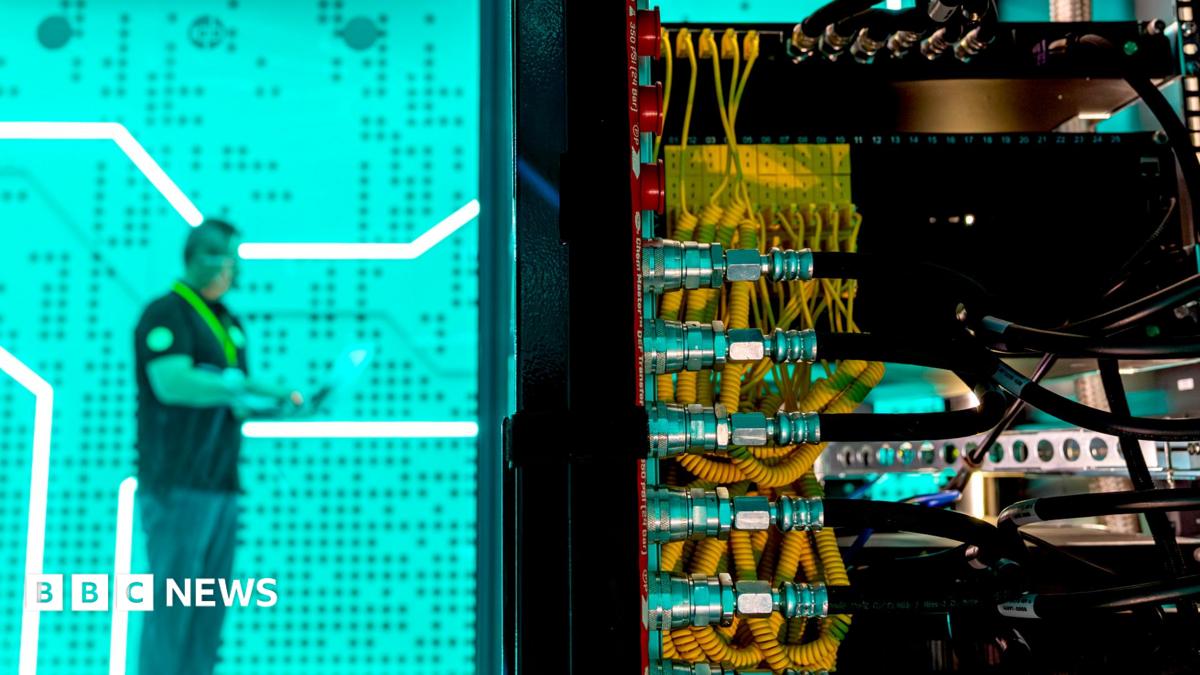 A man standing against a bright green background in a data centre - in the foreground is the back of a large computer with lots of yellow and black wires.