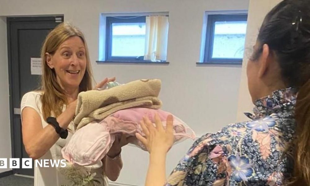 A woman holding a number of items that look like blankets, with a woman handing them to her. They both have long hair.