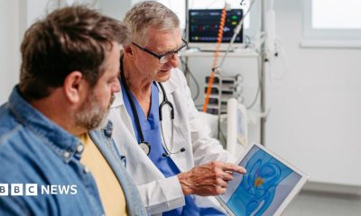 The image shows a clinical setting with medical equipment visible in the background, including a monitor displaying vital signs. In the foreground, a healthcare professional wearing a white coat and a stethoscope is seated next to another adult man. The healthcare professional is pointing to a tablet screen that displays a simplified anatomical illustration highlighting the prostate in orange. The second individual is looking toward the tablet, seated closely and facing the healthcare professional. The environment appears clean and well-lit, with light-coloured walls and organised medical devices.