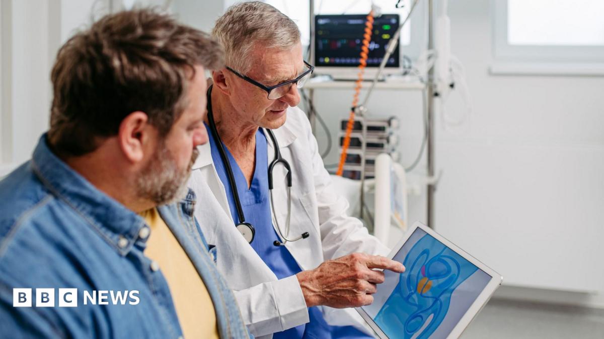 The image shows a clinical setting with medical equipment visible in the background, including a monitor displaying vital signs. In the foreground, a healthcare professional wearing a white coat and a stethoscope is seated next to another adult man. The healthcare professional is pointing to a tablet screen that displays a simplified anatomical illustration highlighting the prostate in orange. The second individual is looking toward the tablet, seated closely and facing the healthcare professional. The environment appears clean and well-lit, with light-coloured walls and organised medical devices.