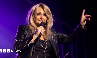 A glamorous woman with blonde hair, wearing all black and holding a microphone, with one hand raised up in the air.
