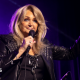 A glamorous woman with blonde hair, wearing all black and holding a microphone, with one hand raised up in the air.