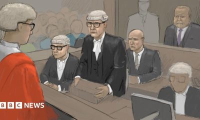 Courtroom sketch showing a legal hearing in progress. A judge in a red robe is seen from behind in the foreground, facing several barristers wearing traditional black robes, white neck bands, and white wigs. One barrister stands at a lectern addressing the court while others are seated nearby. Robert Bush is seated behind a glass partition and other figures are in the public gallery. The image is rendered in a muted palette of grey, brown, and black tones, with the judge’s bright red robe providing a strong visual focal point.
