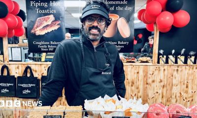 Romesh stood behind a counter of baked goods. He is wearing an apron and black cap and is smiling directly at the camera. There are red and black balloons behind him