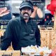 Romesh stood behind a counter of baked goods. He is wearing an apron and black cap and is smiling directly at the camera. There are red and black balloons behind him