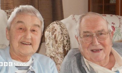 An elderly woman and man smile at the camera as they sit in armchairs. The woman has short grey hair and wears a light blue cardigan and a white top underneath with grey and black stripes on it. The man wears a white shirt with a collar and a grey jumper over the top.