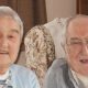 An elderly woman and man smile at the camera as they sit in armchairs. The woman has short grey hair and wears a light blue cardigan and a white top underneath with grey and black stripes on it. The man wears a white shirt with a collar and a grey jumper over the top.